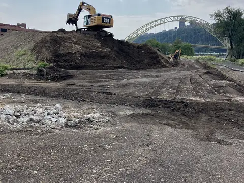 Excavator on a spoil pile with the West End Bridge beyond. Site work, August 2026