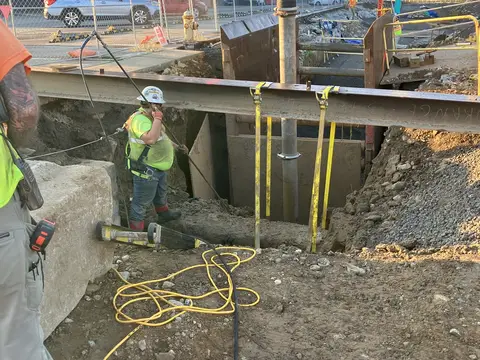 Crew setting a support beam over an excavation. Site work, August 2026